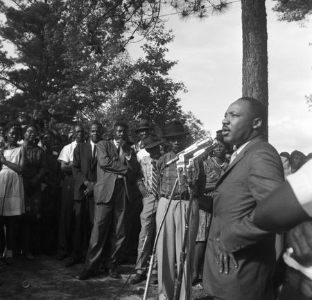 Martin Luther King Jr. addressing an audience at a voter rally in a wooded area in Alabama.